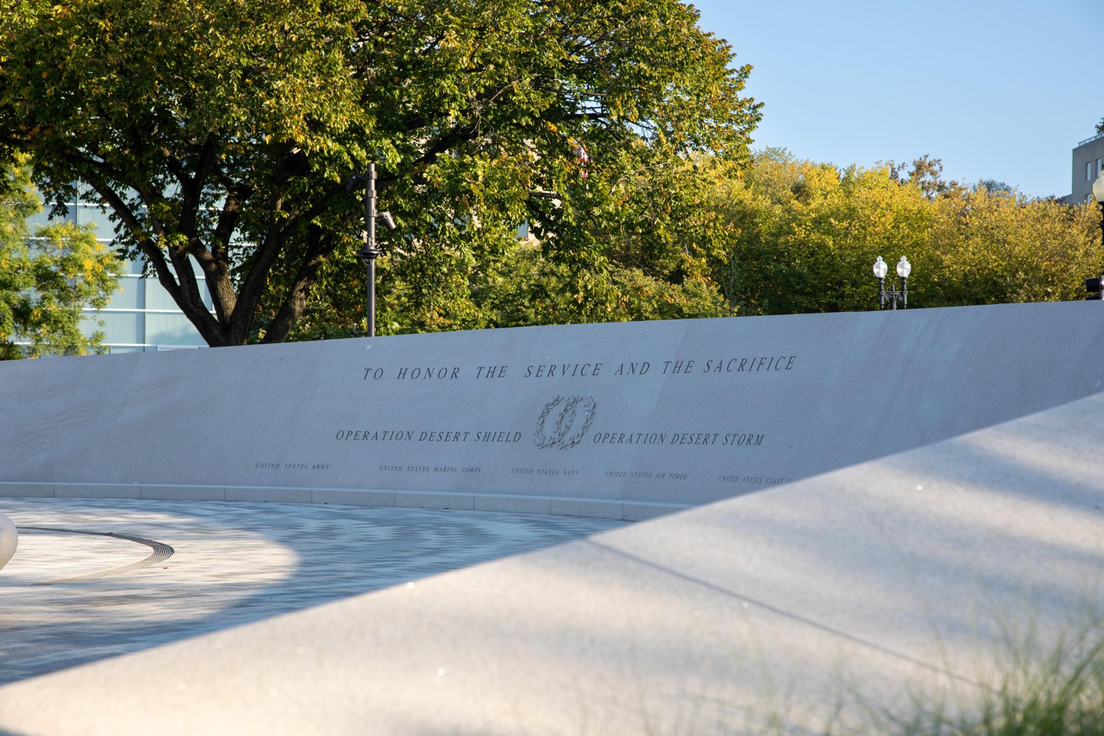 Desert Shield and Desert Storm Memorial: curved stone wall inscribed "To Honor the Service and the Sacrifice," with trees behind it.